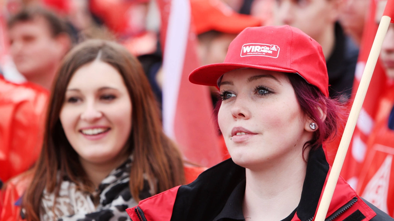 Zwei lächelnde Frauen, Eine trägt eine IG Metall-Kappe und eine Fahne über die Schulter.